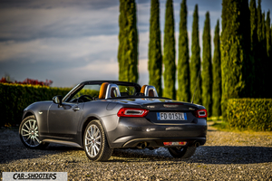 Fiat 124 Spider – rear three-quarter, grey metallic on a gravel driveway with cypress trees