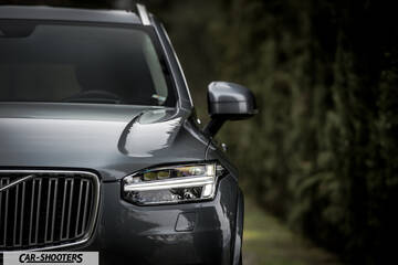 Volvo XC90 – front three-quarter, grey metallic against a blurry hedge backdrop