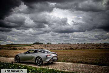 Lexus LC500h – rear three-quarter, silver on a rural road under a stormy sky
