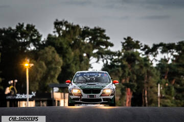 front three-quarter, white with green and orange driving on a track surrounded by trees