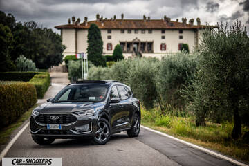 Ford Kuga 2025 – front three-quarter, dark grey on a country road with a large building in the background