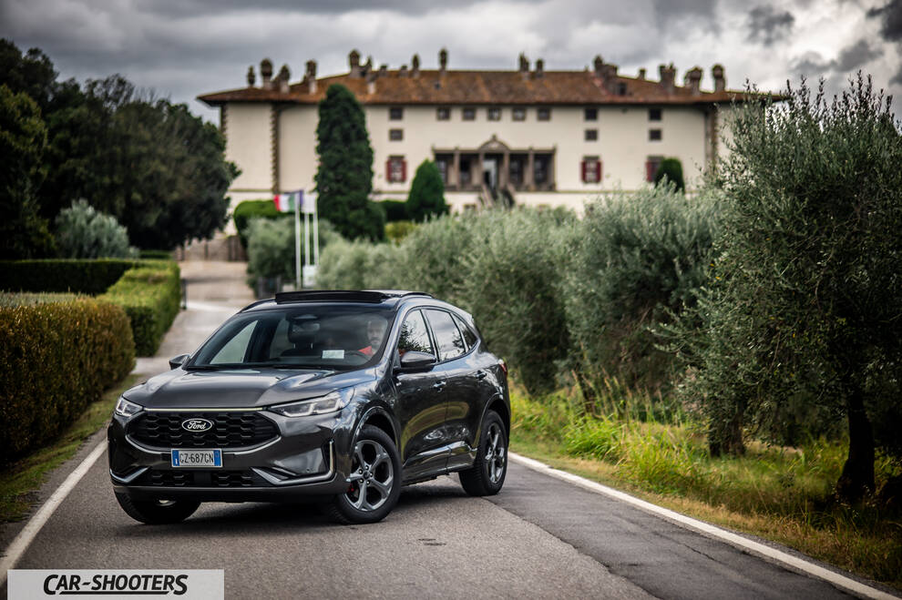 Ford Kuga 2025 – front three-quarter, dark grey on a country road with a large building in the background